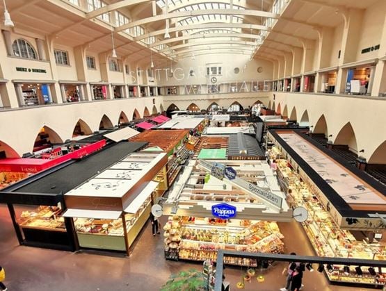 Vue intérieure de la Markthalle de Stuttgart, un grand marché couvert baigné de lumière naturelle. Sous une haute verrière, de nombreux stands colorés proposent une variété de produits alimentaires et spécialités locales, dans une ambiance conviviale et animée.