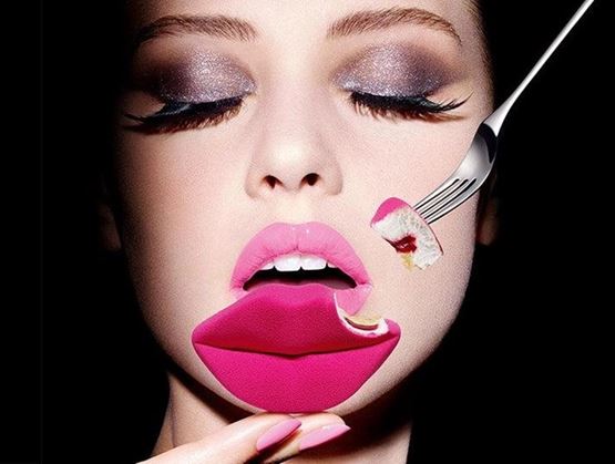 Image artistique d’une femme maquillée de manière glamour, avec des paupières scintillantes et des lèvres rose vif stylisées en forme de gâteau. Une fourchette s’approche de sa bouche avec une bouchée de dessert, créant une mise en scène audacieuse mêlant beauté et gourmandise.