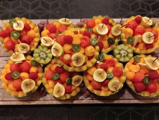 Assortiment de salades de fruits frais joliment dressées dans des coques de melon évidées. Chaque composition contient des billes de melon et de pastèque, des tranches de banane, de kiwi, des framboises et des mini-kumis, formant un ensemble coloré et appétissant sur une planche en bois rustique.