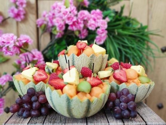 Saladiers naturels confectionnés à partir de melons évidés, remplis de morceaux de fruits frais colorés tels que fraises, kiwi, ananas, framboises et billes de melon. Le tout est joliment présenté sur une table en bois, avec des grappes de raisin et un fond fleuri décoratif.