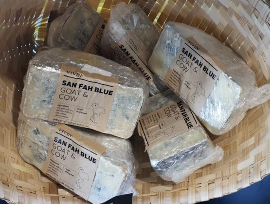 Panier en osier contenant plusieurs portions de fromage bleu « San Fah Blue Goat & Cow » emballées dans du film plastique. Les étiquettes précisent qu’il s’agit d’un produit artisanal, local et naturel à base de lait de chèvre et de vache.