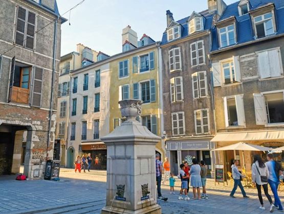 Place de centre-ville animée à Pau, avec une fontaine en pierre au premier plan. Des passants circulent entre les façades colorées et les terrasses de café, créant une ambiance conviviale et ensoleillée.