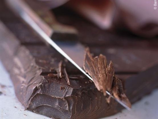 Gros plan sur une tablette de chocolat noir en train d’être découpée avec un couteau, faisant apparaître des éclats et copeaux fins. L’image met en valeur la texture fondante et la qualité artisanale du chocolat.