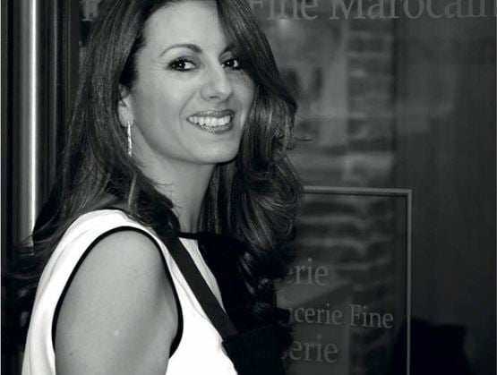 Portrait en noir et blanc d’une femme souriante aux cheveux ondulés, habillée avec élégance. Elle se tient devant une vitrine sur laquelle on distingue partiellement les mots « Épicerie Fine ». L’image évoque un cadre professionnel et raffiné.