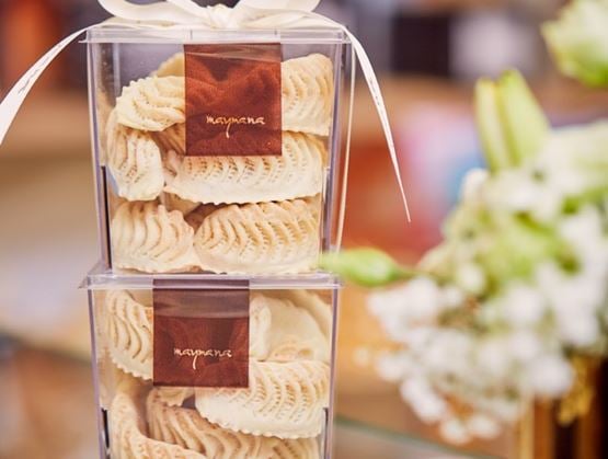 Deux boîtes transparentes empilées, remplies de pâtisseries fines blanches ornées de motifs délicats, avec une étiquette « Maymana » sur chacune. L’ensemble est décoré d’un ruban, suggérant un coffret cadeau raffiné et artisanal.