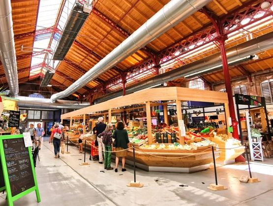 Vue intérieure d’un marché couvert à Colmar, avec une architecture mêlant bois, métal rouge et verrière. Au centre, un grand étal de fruits et légumes attire les clients dans une ambiance lumineuse et conviviale.