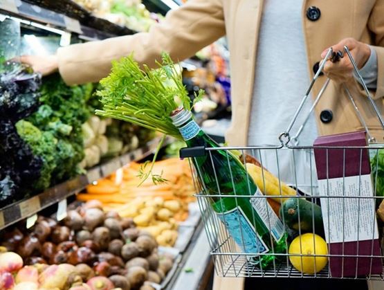 Personne faisant ses courses dans un rayon de fruits et légumes frais, tenant un panier rempli de produits sains : eau minérale, citron, avocat, céleri et emballages alimentaires. L’ambiance évoque un mode de vie urbain et attentif à l’alimentation.