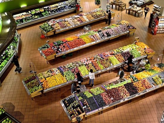 Vue en plongée d’un rayon fruits et légumes dans un supermarché, avec de longs étals colorés remplis de produits frais. Plusieurs clients font leurs courses, créant une scène vivante et ordonnée au cœur d’un espace bien éclairé.