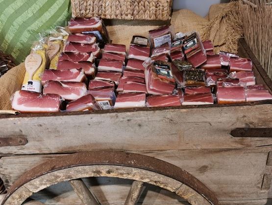 Charrette rustique remplie de morceaux de speck du Tyrol du Sud emballés sous vide, présentés sur un lit de toile de jute dans un décor d’épicerie fine ou de marché artisanal.