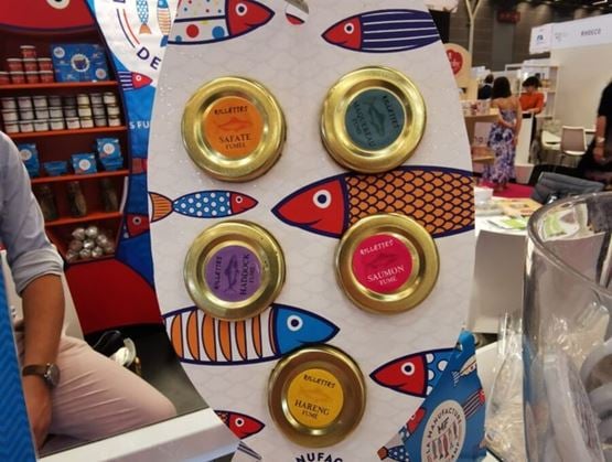 Présentation de six bocaux de rillettes artisanales (safate fumée, lieu, haddock, saumon, maquereau et hareng fumé) sur un présentoir en forme de poisson, aux couleurs vives, au salon Gourmet Sélection.