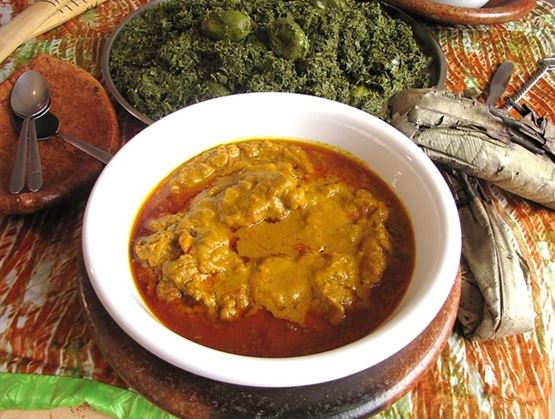 Plat congolais de moambe au poulet servi dans un bol blanc, accompagné de feuilles de manioc et de petits fruits verts sur une assiette à l’arrière-plan, posés sur une nappe colorée.