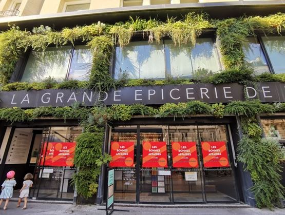 Façade de La Grande Épicerie avec murs végétalisés et affiches rouges annonçant des « Bonnes affaires » en vitrine, deux enfants marchant sur le trottoir au premier plan.