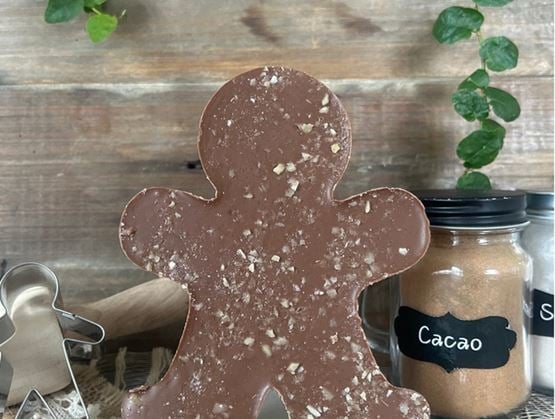Un grand chocolat en forme de bonhomme, parsemé d’éclats, est posé sur une table en bois rustique, entouré d’ustensiles de pâtisserie et de bocaux étiquetés « Cacao » et « Sucre », avec des feuilles vertes en arrière-plan.