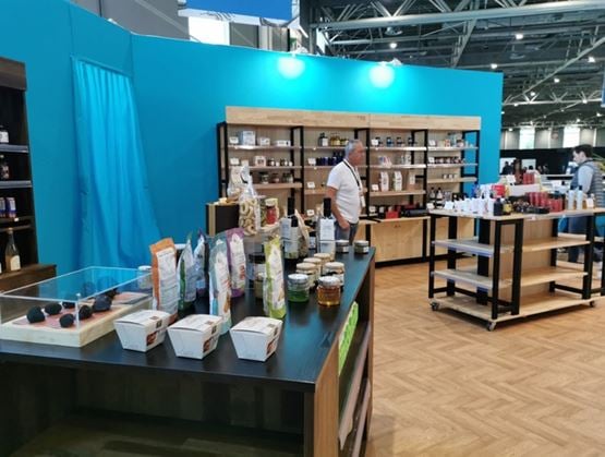 Un stand d’exposition au design moderne avec des murs turquoise présente des produits d’épicerie fine disposés sur des tables et des étagères en bois, tandis qu’un exposant se tient derrière le comptoir dans un hall de salon professionnel.