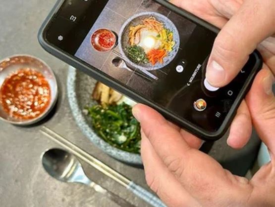 Une personne photographie avec son smartphone un bol de plat coloré présenté sur une table, accompagné de petits bols de sauce et de couverts disposés à côté.