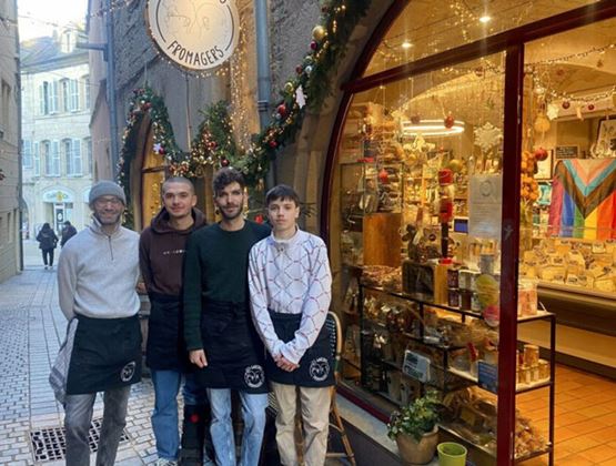 Quatre fromagers posent devant la devanture décorée de leur boutique artisanale pendant les fêtes, avec vitrines remplies de produits du terroir et décorations de Noël scintillantes.