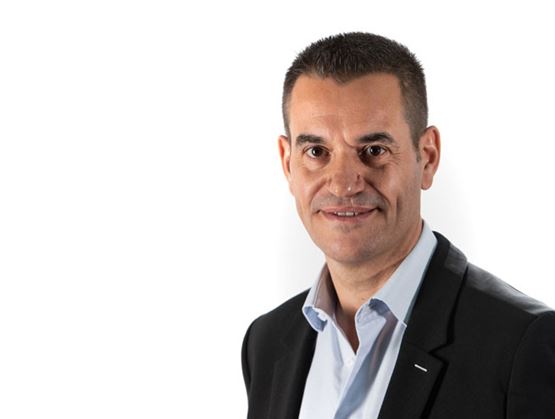 Portrait d’un homme en veste noire et chemise claire, souriant, sur fond blanc.