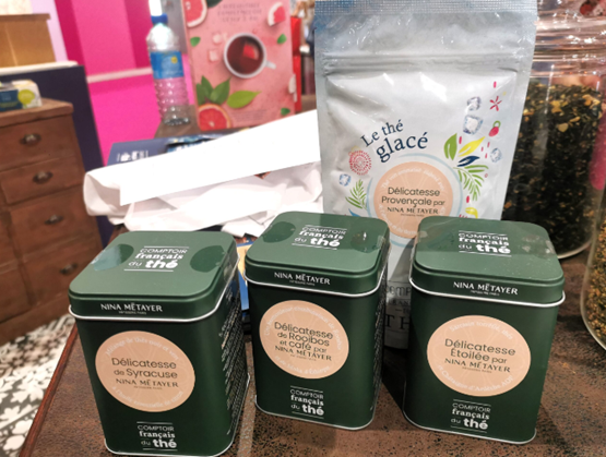 "Boîtes de thé vertes de la collection Nina Métayer du Comptoir Français du Thé, exposées sur un stand, avec en arrière-plan un sachet de thé glacé aux motifs floraux."