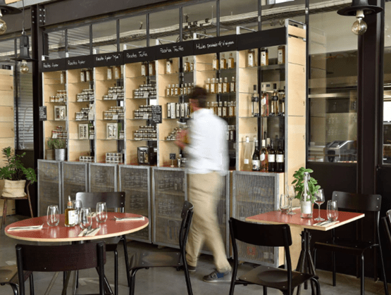 Épicerie fine intégrée à un restaurant contemporain en Provence, imaginée par le chef Julien Allano. On y voit des étagères élégantes garnies de produits artisanaux : huiles, condiments, truffes et alcools fins. Un homme en mouvement parcourt les rayons, tandis que des tables dressées accueillent les clients dans une ambiance conviviale et moderne.