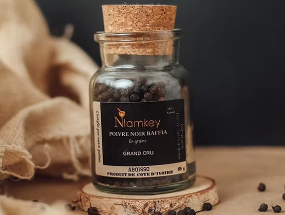 Bocal en verre transparent contenant du poivre noir Baffia en grains de la marque Niamkey, fermé par un bouchon en liège. L’étiquette noire indique "Grand Cru", "Produit de Côte d’Ivoire", et précise son origine : Abidjan, Aboisso. Le bocal est posé sur une tranche de bois, avec quelques grains de poivre dispersés autour, dans une mise en scène naturelle et artisanale. L’image évoque un produit d’exception issu d’un terroir africain valorisé.