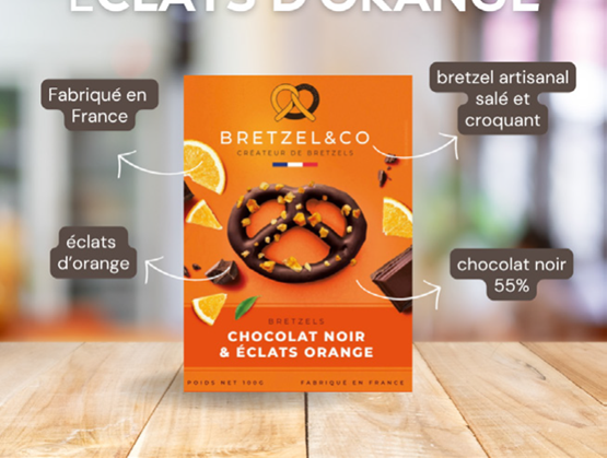 Packaging du produit "Bretzel&Co – Chocolat noir & éclats orange", présenté au centre sur un fond en bois clair. Le visuel met en avant un bretzel enrobé de chocolat noir 55 %, parsemé d’éclats d’orange. Des annotations signalent les atouts du produit : fabriqué en France, bretzel artisanal salé et croquant, éclats d’orange, et chocolat noir 55 %. L’ensemble évoque une gourmandise artisanale mêlant tradition et originalité.