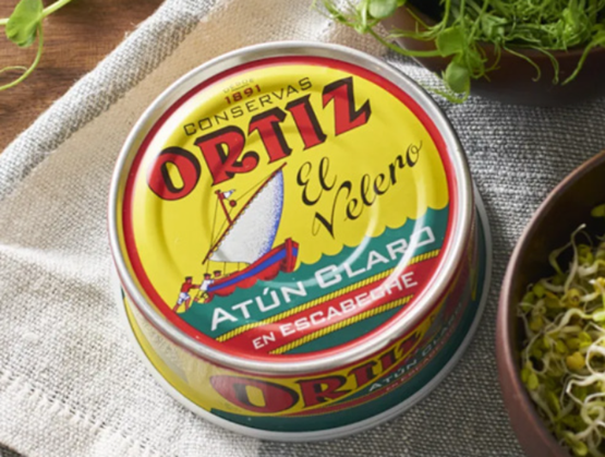 ChatGPT said: Colorful box of white tuna marinated in escabeche from the brand Conservas Ortiz, placed on a beige cloth with a wooden background. The vintage label, dominated by yellow, red, and blue, features an illustration of a traditional fishing boat. Around the box, fresh herbs and sprouts suggest a natural and refined culinary world.