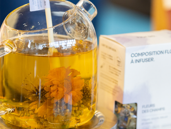 Théière en verre contenant une infusion florale avec des fleurs entières visibles, notamment une grande fleur orange. À côté, une boîte de la marque Horae mentionnant "Composition florale à infuser – Fleurs des champs". L’image met en valeur une infusion artisanale et poétique, issue de fleurs séchées, dans un cadre lumineux et apaisant.