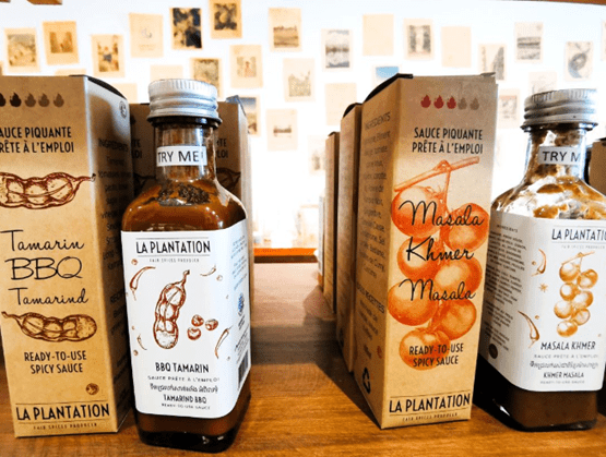 Flacons de sauces épicées "La Plantation" exposés sur un comptoir en bois, accompagnés de leurs emballages. À gauche, une sauce BBQ au tamarin, à droite une sauce Masala Khmer. Les packagings mettent en avant des illustrations d’ingrédients naturels et des mentions en français et en anglais. En arrière-plan, un mur décoré de cartes ou illustrations anciennes. 