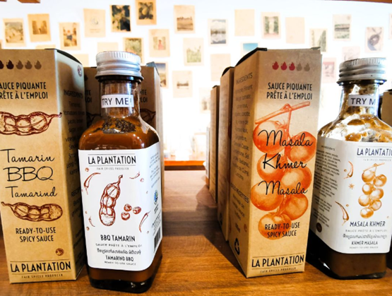 Flacons de sauces épicées "La Plantation" exposés sur un comptoir en bois, accompagnés de leurs emballages. À gauche, une sauce BBQ au tamarin, à droite une sauce Masala Khmer. Les packagings mettent en avant des illustrations d’ingrédients naturels et des mentions en français et en anglais. En arrière-plan, un mur décoré de cartes ou illustrations anciennes.