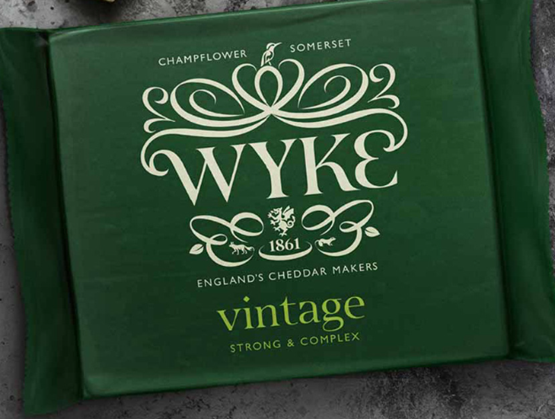 Emballage vert foncé d’un cheddar "Vintage" de la marque Wyke Farms, originaire de Champflower dans le Somerset (Angleterre). Le design raffiné met en avant le nom "WYKE" avec des ornements blancs élégants, ainsi que la mention "England’s Cheddar Makers – Since 1861". L’inscription "Vintage – Strong & Complex" indique un fromage affiné à la saveur prononcée et complexe. Le packaging évoque tradition, savoir-faire et qualité artisanale britannique.