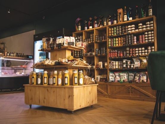 Vue intérieure d’une épicerie fine chaleureuse, avec un sol en bois clair et des étagères en bois remplies de produits artisanaux : bocaux, conserves, pâtes, huiles et confiseries. Au centre, un présentoir en bois expose des bouteilles de jus, du pain frais et des produits locaux. L’ambiance est cosy et authentique, évoquant le terroir et la qualité.