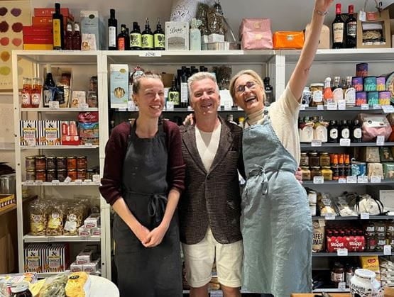 Trois personnes souriantes, dont deux en tablier, posent devant les étagères remplies de produits fins dans une épicerie chaleureuse. Ambiance conviviale et esprit de quartier au cœur de l’épicerie June.
