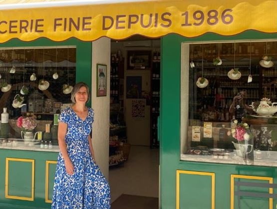 Femme souriante en robe bleue devant une épicerie fine traditionnelle à la devanture verte et jaune, avec une enseigne indiquant “Épicerie fine depuis 1986”. Vitrine décorée de produits et de tasses suspendues.