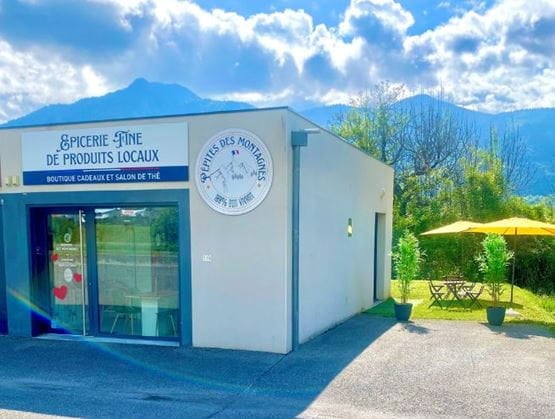 Façade moderne de l’épicerie “Pépites des Montagnes”, spécialisée en produits locaux, avec vue sur les montagnes. À droite, un coin détente extérieur avec parasol jaune et table de jardin.