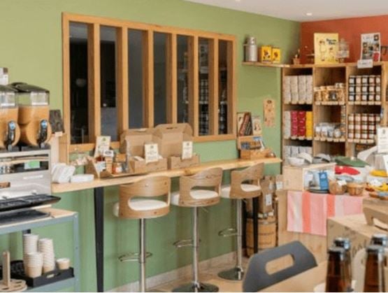 Intérieur chaleureux d’une épicerie fine avec comptoir en bois, tabourets hauts, produits artisanaux exposés et coin café. Ambiance conviviale et décor soigné mêlant bois clair et mur vert pastel.
