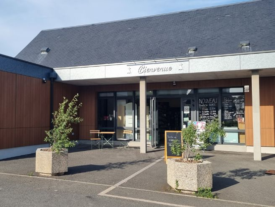 Entrance of the “Au Petit Marché” grocery store in La Gouesnière, featuring a modern façade, potted plants, a welcome chalkboard, and a terrace with tables