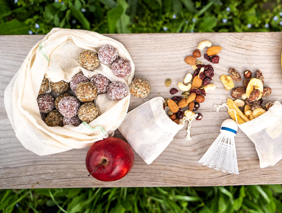 Healthy snacks arranged on a wooden board outdoors, with energy balls in a cloth bag, dried fruits and nuts, a red apple, and a badminton shuttlecock.