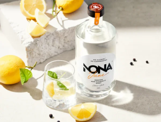 Bouteille de Nona June, un spiritueux sans alcool, posée sur une surface claire avec un verre garni de citron, basilic et glaçons. Des citrons frais et des baies de genièvre décorent la scène en lumière naturelle.
