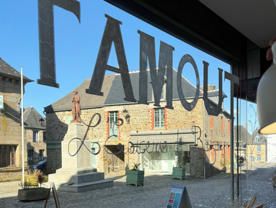 Vue depuis l’intérieur de l’épicerie "Tamout, L’Épicerie de Bâzouj" à Bazouges-la-Pérouse, montrant la place du Monument et une statue en pierre.