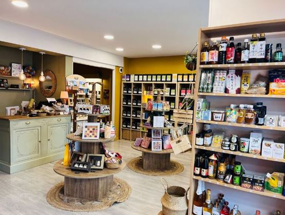 Vue intérieure d’une boutique spécialisée, chaleureusement aménagée, avec des étagères remplies de produits artisanaux, d’épicerie fine, de thés et de boissons. Au centre, des présentoirs en bois exposent des articles soigneusement disposés. Le comptoir d’accueil se trouve à gauche, avec une décoration cosy et des luminaires suspendus.