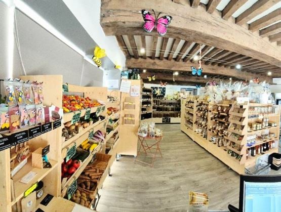 Intérieur d’une épicerie fine à l’ambiance rustique, avec un plafond en poutres apparentes et une décoration colorée comprenant des papillons suspendus. Des étagères en bois présentent des fruits et légumes frais à gauche, ainsi que des produits artisanaux, bocaux et paniers garnis à droite.