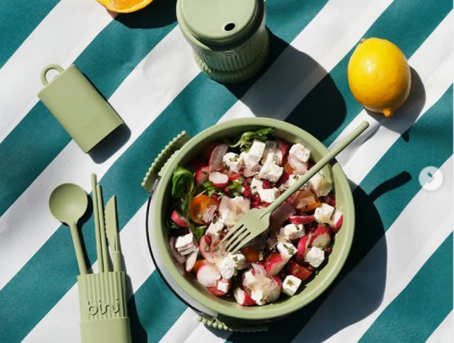Green reusable Bini picnic set with matching cutlery, lunch box, and cup, placed on a striped tablecloth alongside a fresh salad, a lemon, and half an orange.