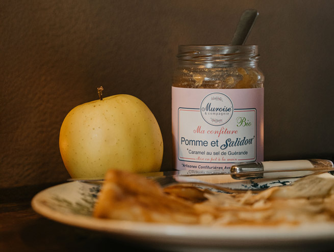 Pot de confiture Pomme et Salidou® de Muroise et Compagnie, posé à côté d’une pomme jaune, avec une assiette de crêpes au premier plan.