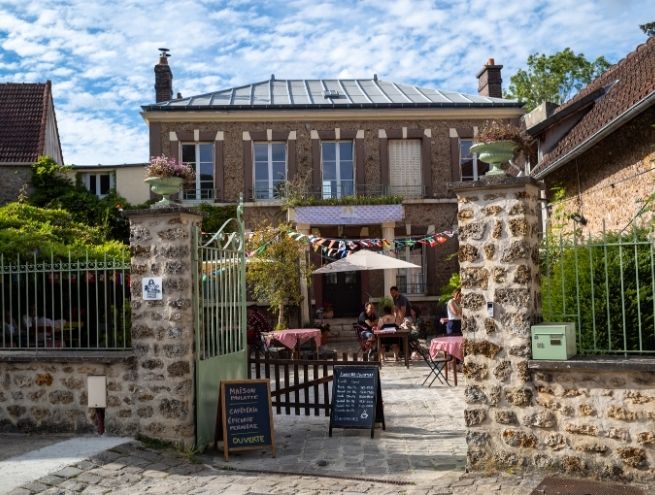 Façade d’une maison-épicerie de campagne avec terrasse, tables extérieures et ambiance conviviale.