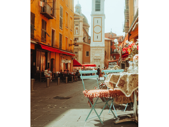 Terrasse de café dans une rue pavée d’une ville méditerranéenne, avec une table dressée au premier plan, des chaises pliantes colorées, des façades aux tons chauds et un clocher en arrière-plan.