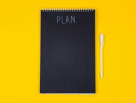 Carnet noir sur lequel est incrit notes en blanc avec un stylo blanc posé à sa droite sur fond jaune