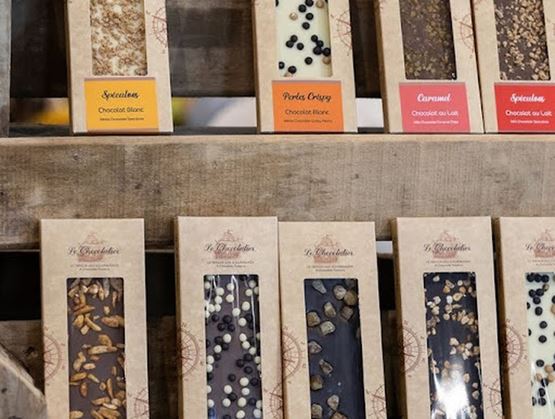 Tablette de chocolats artisanaux présentés dans des emballages en carton, disposés sur des étagères en bois, avec différentes saveurs et garnitures visibles comme des fruits secs, des perles et des épices.