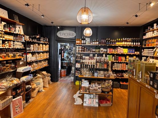 Intérieur d’une épicerie fine chaleureuse avec des étagères en bois remplies de produits artisanaux, bocaux, bouteilles et paniers, éclairée par des suspensions décoratives.