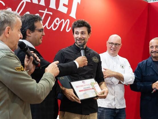 Un lauréat tient un certificat et s’exprime micro en main sur la scène de Gourmet Selection, entouré de plusieurs intervenants devant un fond rouge." (Default Alternate Text: "Un lauréat tient un certificat et s’exprime micro en main sur la scène de Gourmet Selection, entouré de plusieurs intervenants devant un fond rouge.