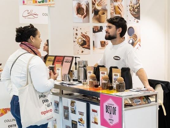 Un exposant échange avec une visiteuse sur un stand BIBO lors d’un salon, devant une présentation de boissons, d’échantillons et de visuels produits." (Default Alternate Text: "Un exposant échange avec une visiteuse sur un stand BIBO lors d’un salon, devant une présentation de boissons, d’échantillons et de visuels produits.